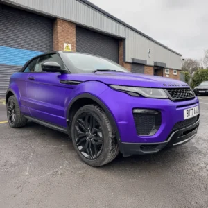 A Range Rover Evoque Convertible wrapped in a satin purple finish, parked outside an industrial unit with black roller shutters. The vehicle features black alloy wheels, a black front grille, and a black soft-top roof, creating a striking contrast. The wrap is smooth and professionally applied, giving the car a sleek and modern look.