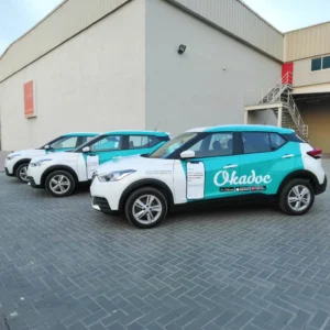 A fleet of three white and turquoise SUVs featuring a professional commercial vehicle wrap for Okadoc, a healthcare app. The branding includes the Okadoc logo in white on the turquoise background, with promotional text on the doors. The vehicles are parked outside an industrial building with a grey brick pavement, a beige warehouse, and a red staircase leading to an upper-level entrance.