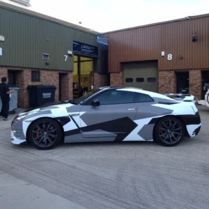 A Nissan GT-R wrapped in a custom black, white, and grey geometric camouflage design, parked outside an industrial unit with brown and green metal cladding. The wrap features sharp angular patterns, giving the car an aggressive and modern appearance. The vehicle has dark alloy wheels with orange brake calipers, tinted windows, and a large rear spoiler. A person is seen in the background near industrial bins, while the garage doors and signage indicate a workshop setting.