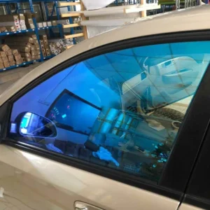 A close-up of a beige car with newly installed blue reflective window tint on the side window. The tint creates a mirrored effect, reflecting the workshop environment, including shelves stocked with rolls of vinyl and tools. The tinted side mirror matches the window, giving a cohesive aesthetic. The reflection of the ceiling lights and surroundings highlights the smooth and professional application of the film.