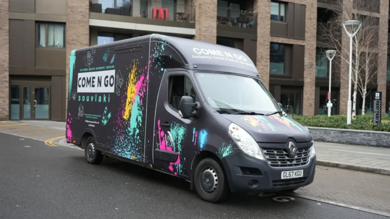 Front view of the Comen Go Souvlaki food truck featuring a vibrant vinyl wrap with splashes of pink, yellow, and blue on a black background, parked on a city street.
