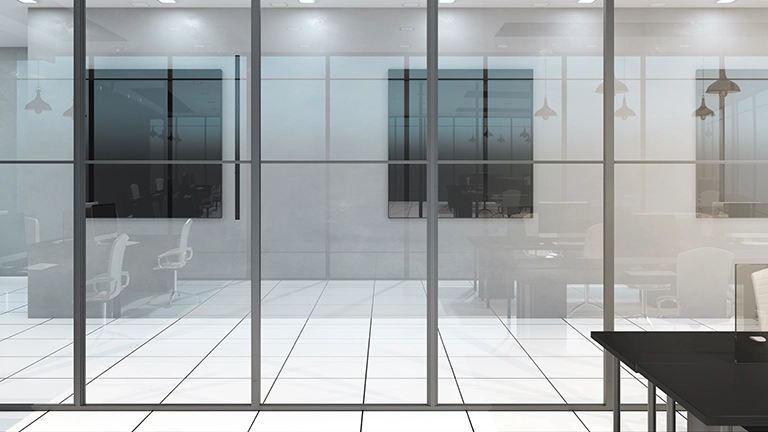 Interior view of a modern office space separated by clear glass partitions, showing desks, white chairs, and ceiling lights, with a reflective white tile floor.
