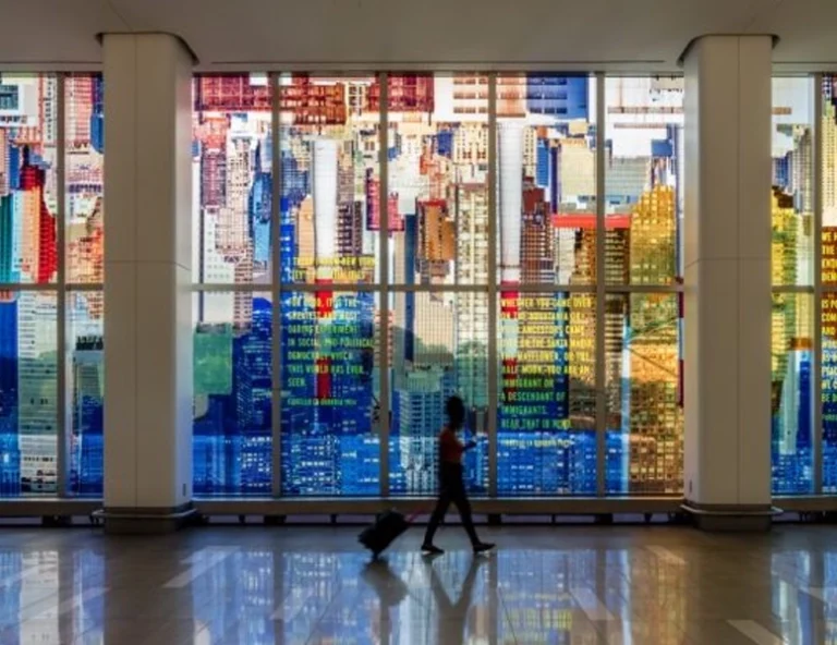A traveler walks past a massive floor-to-ceiling glass wall at LaGuardia Airport, featuring an upside-down colorful cityscape mural with embedded poetic text