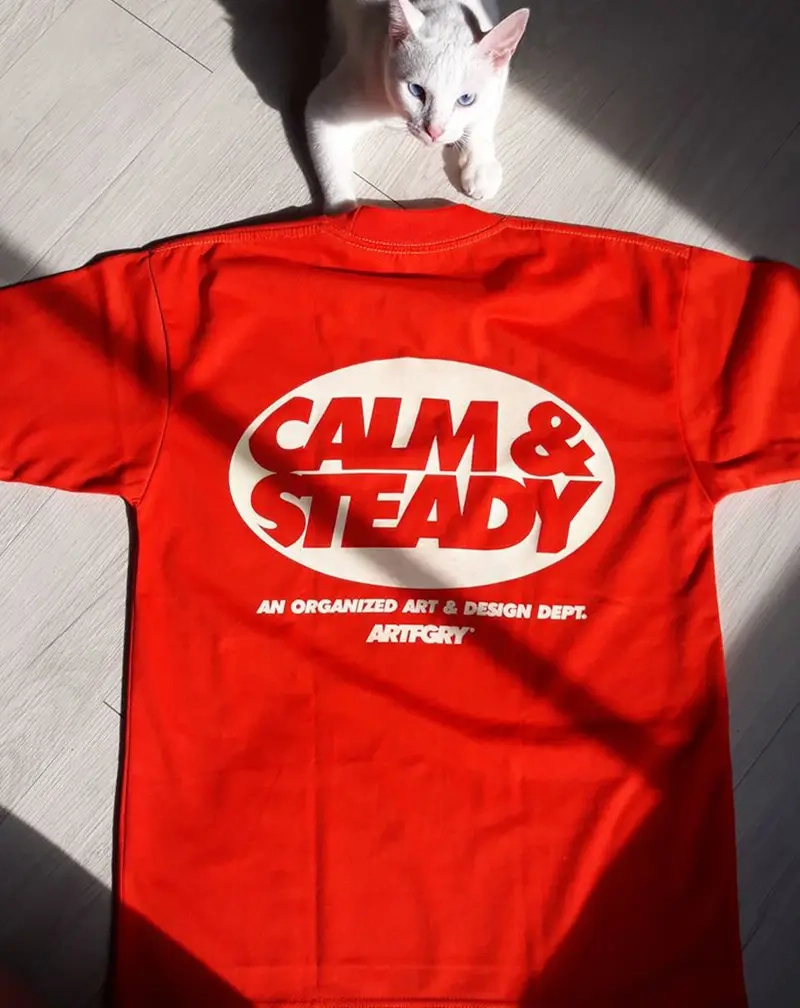 High-angle view of a bright red t-shirt laid on a light wood floor featuring a large cream-colored oval graphic with the text "CALM & STEADY".