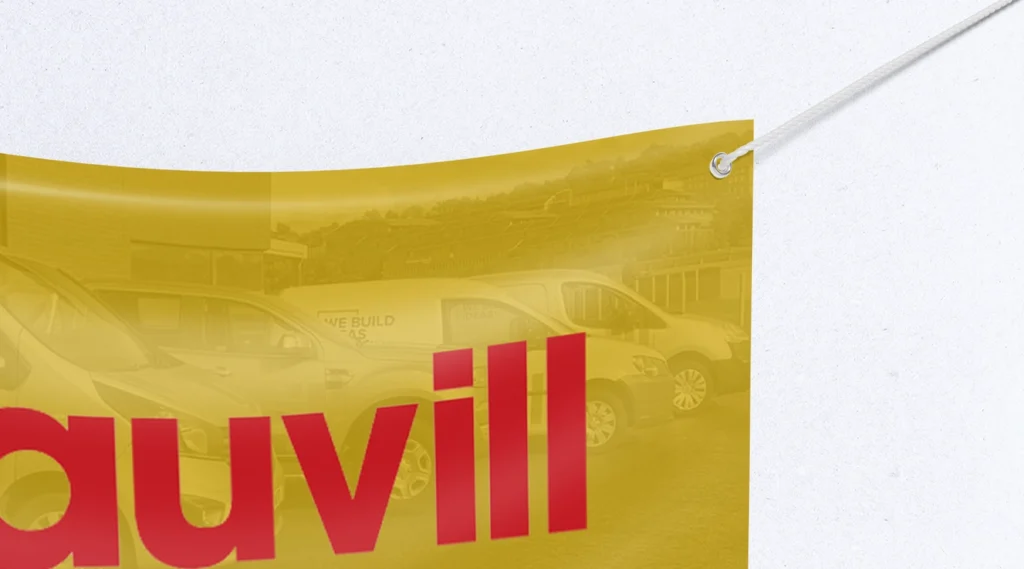 A close-up of a yellow PVC banner for 'Sauvill' featuring red typography and a background image of branded white vans, with a white rope passing through a silver eyelet