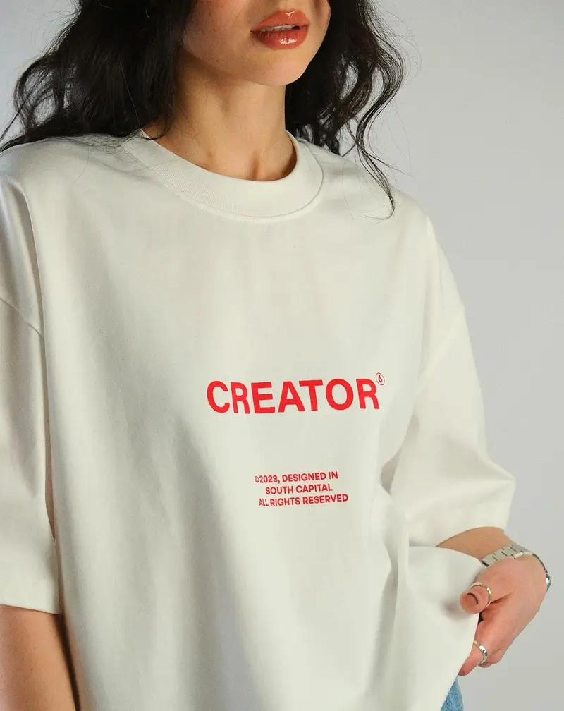 Front view of a person wearing a white heavyweight cotton t-shirt featuring the word "CREATOR" printed in bold red sans-serif typography.