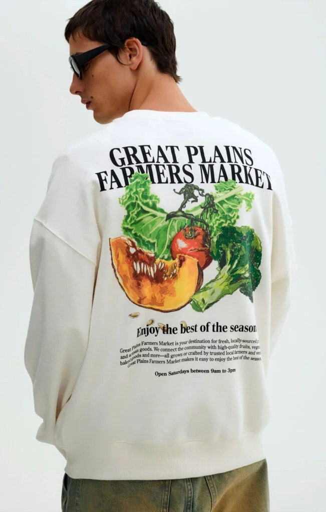 ear view of a person wearing an oversized cream sweatshirt with large-scale "GREAT PLAINS FARMERS MARKET" vegetable graphics printed on the back.