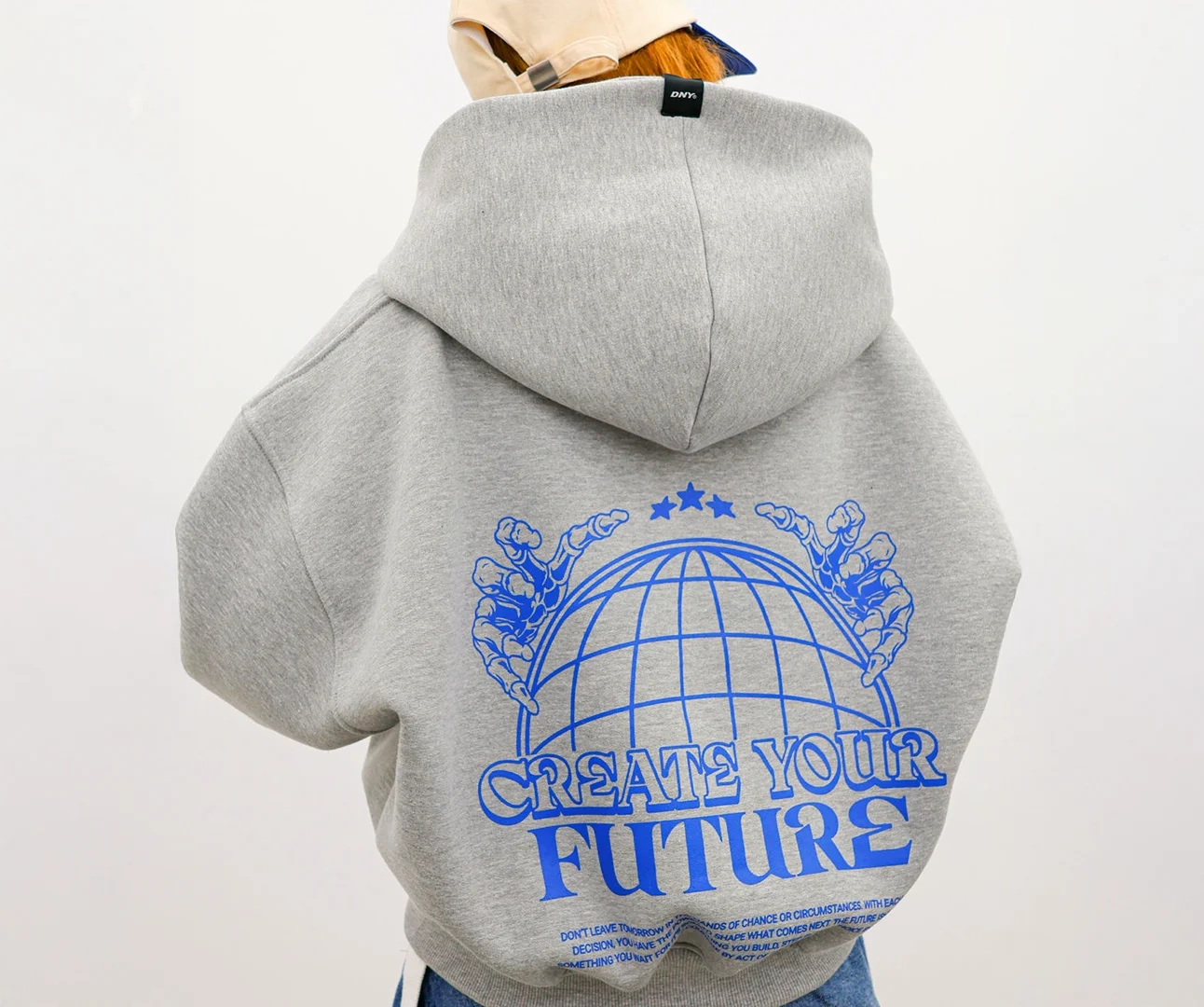 Rear view of a person wearing an oversized heather grey hoodie featuring a large blue graphic of a globe held by skeletal hands with the text "CREATE YOUR FUTURE".