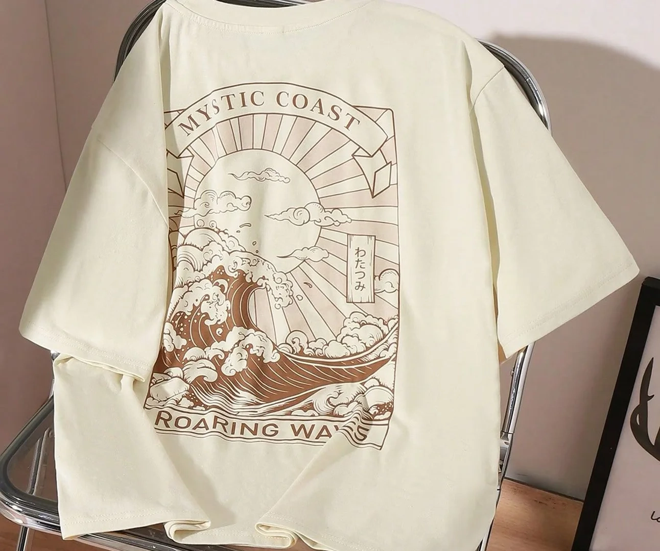 Rear view of a cream-colored t-shirt draped over a chair, featuring a large brown rectangular graphic of a sun and crashing waves with the text "MYSTIC COAST" and "ROARING WAVES".