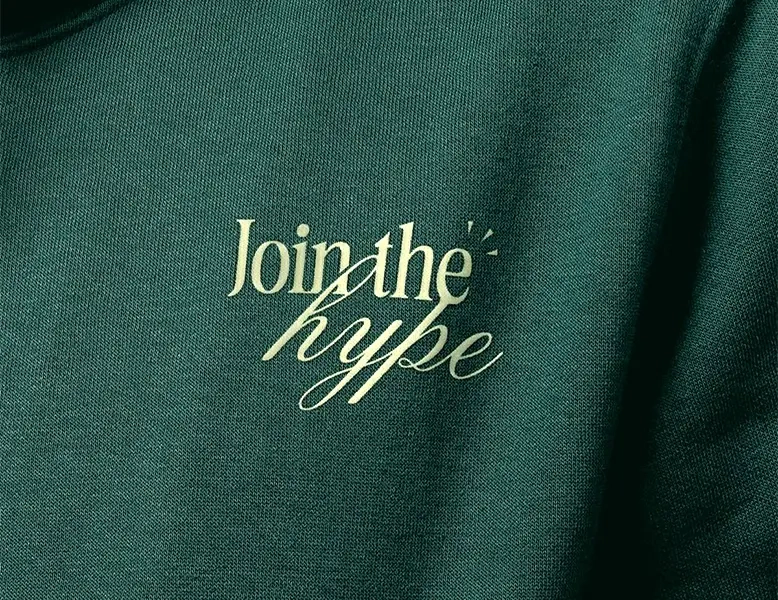 Close-up of a dark green sweatshirt featuring a "Join the hype" graphic printed in a pale cream elegant font.