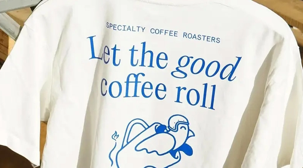 Rear view of a white t-shirt hanging on a white hanger, featuring blue screen-printed graphics including a "bird in a sidecar" illustration and coffee-related text.