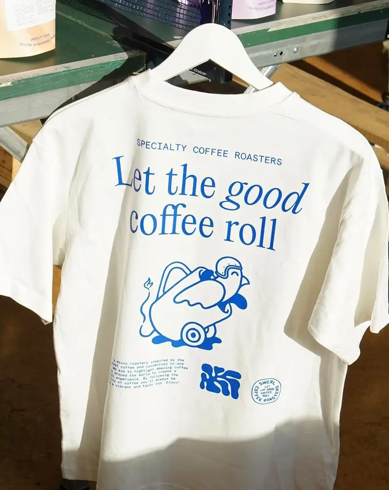 Rear view of a white t-shirt hanging on a white hanger, featuring blue screen-printed graphics including a "bird in a sidecar" illustration and coffee-related text.