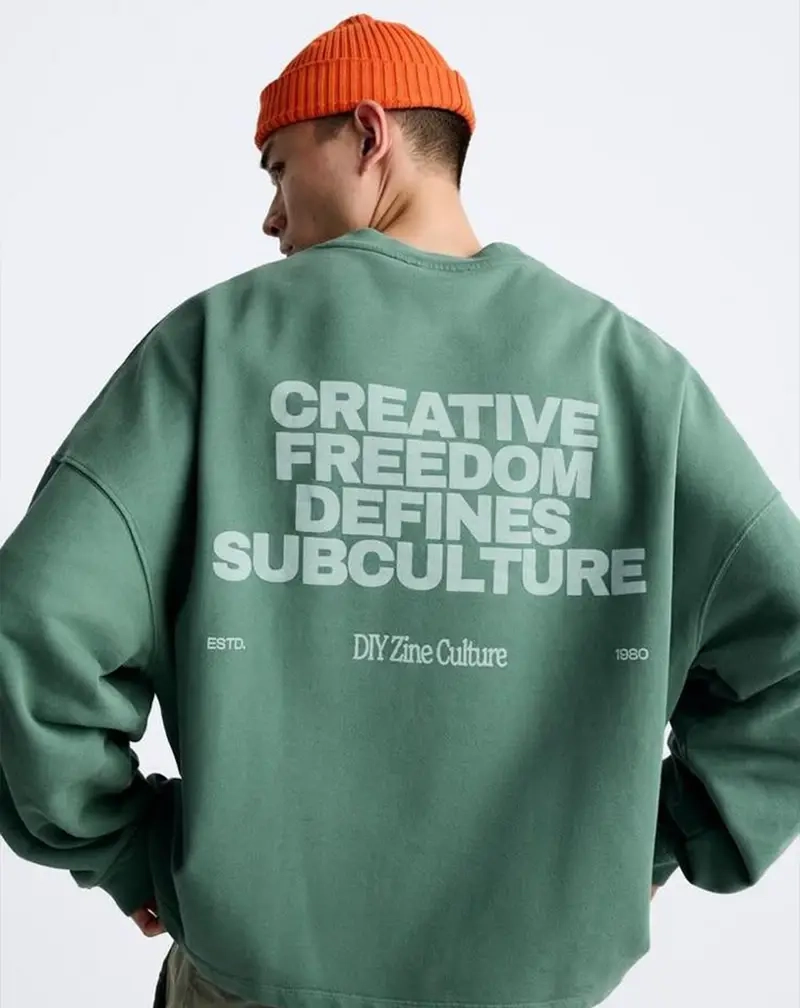 Rear view of a person wearing an oversized sage green sweatshirt with bold light grey typography that reads "CREATIVE FREEDOM DEFINES SUBCULTURE".