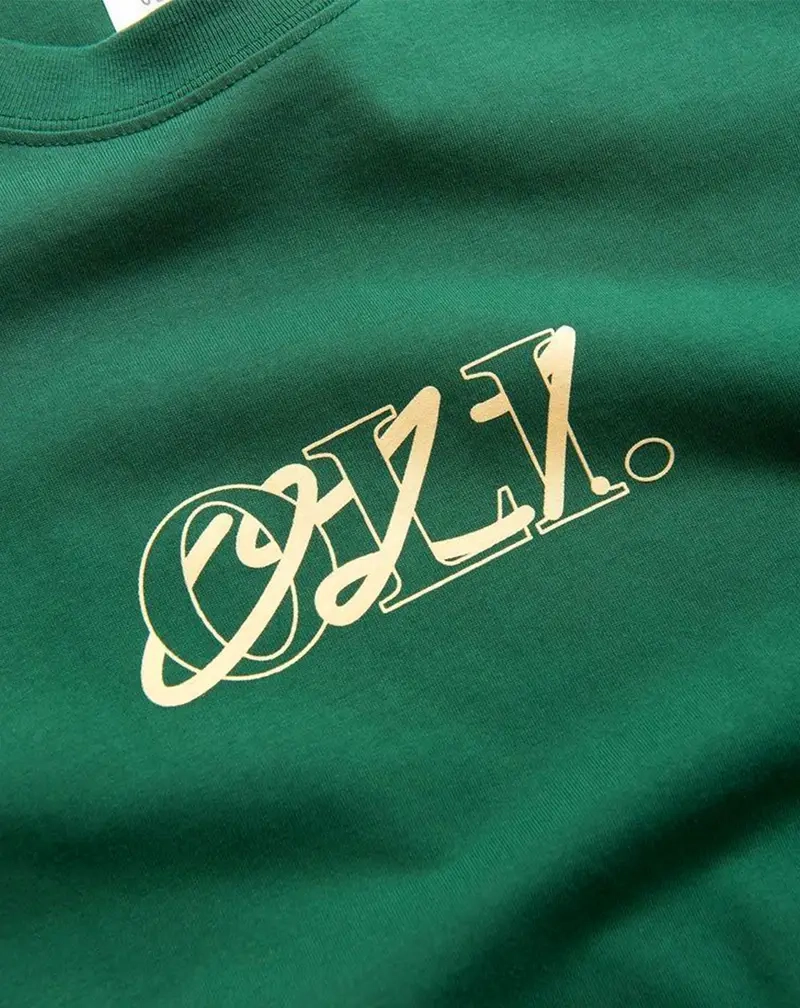 Close-up of a green t-shirt or sweatshirt featuring a stylized script logo "O.Z.X." printed in a pale gold or cream color.