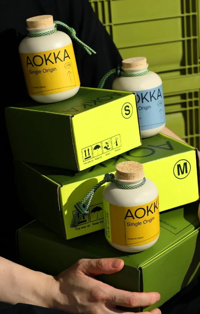 Minimalist white coffee canisters with yellow and blue "AOKKA Single Origin" labels, held alongside lime green shipping boxes with black industrial iconography
