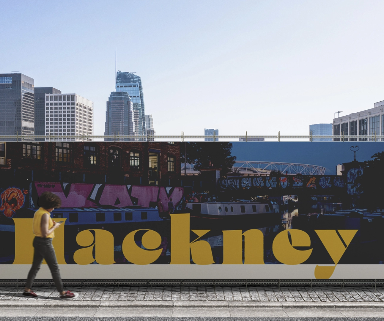 Large-scale outdoor site hoarding along a city sidewalk featuring a panoramic night photograph of a canal with graffiti-covered buildings and bold yellow "Hackney" typography.