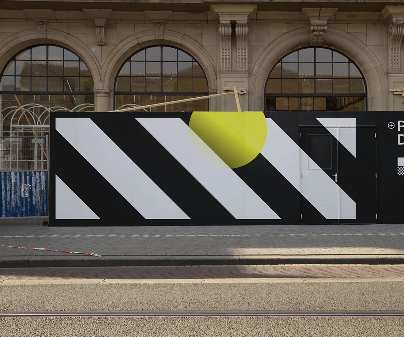 Large-format construction site hoarding panels with black and white diagonal stripes and a yellow circle graphic in front of a historic building.