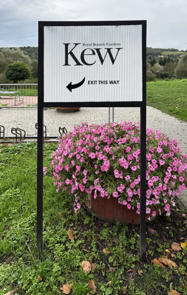 A white fluted corex wayfinding sign for the Royal Botanic Gardens Kew, mounted on a black metal frame in an outdoor garden setting with pink flowers.