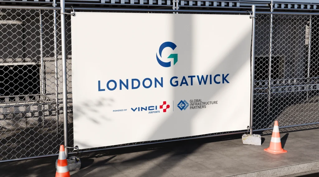 A white vinyl banner for "LONDON GATWICK" secured to a chain-link fence at a construction site, featuring corporate logos and safety cones.