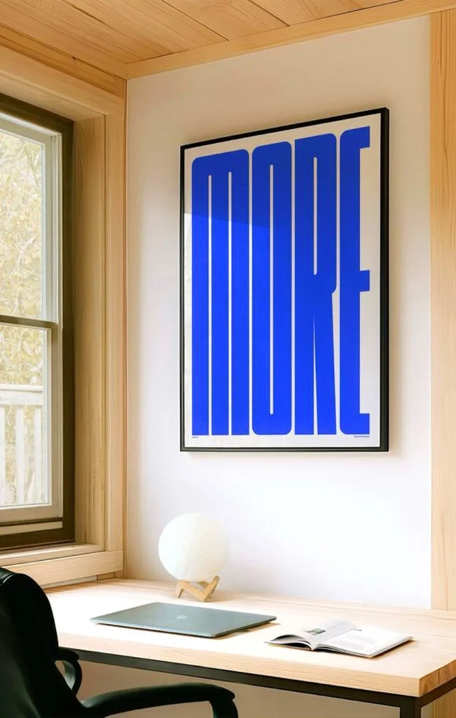 A large, framed indoor wall poster with bold blue "MORE" typography displayed in a minimalist home office setting.