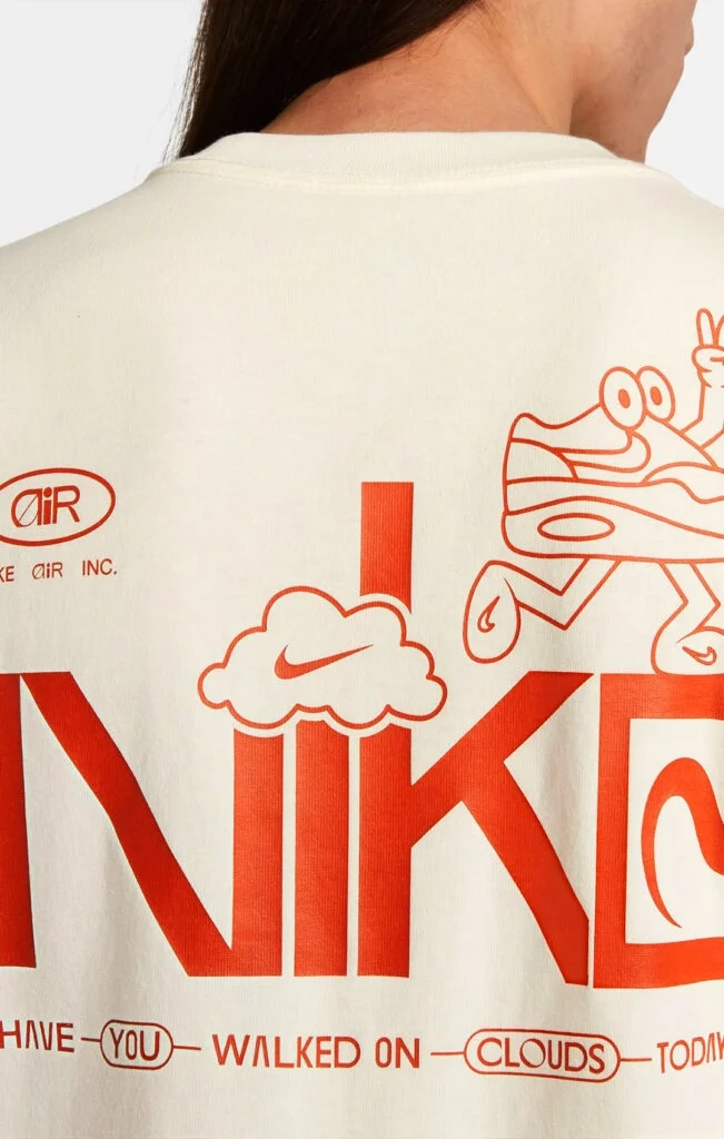 Rear close-up of a cream-colored Nike t-shirt featuring vibrant orange screen-printed graphics including a "walking shoe" character and a cloud logo.