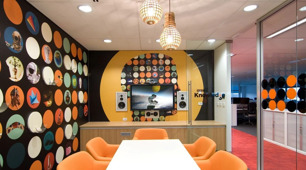 A modern office meeting room featuring a large black and yellow wall graphic of a human head silhouette filled with colorful circular icons, branded with the "BBC Knowledge" logo.