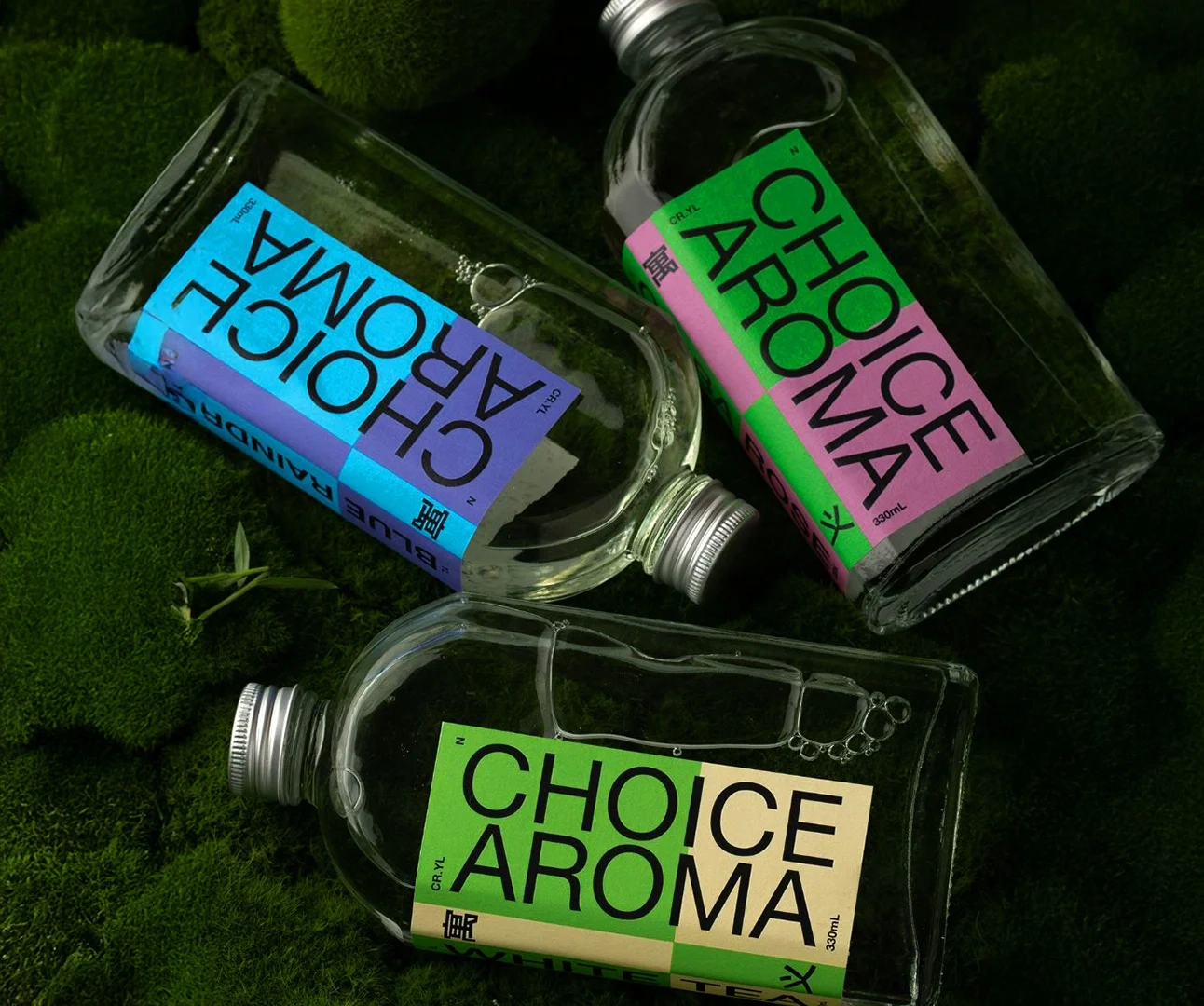 Three clear glass bottles of Choice Aroma specialty drinks with vibrant, color-blocked labels in blue/purple, green/pink, and tan/green, resting on a bed of green moss.