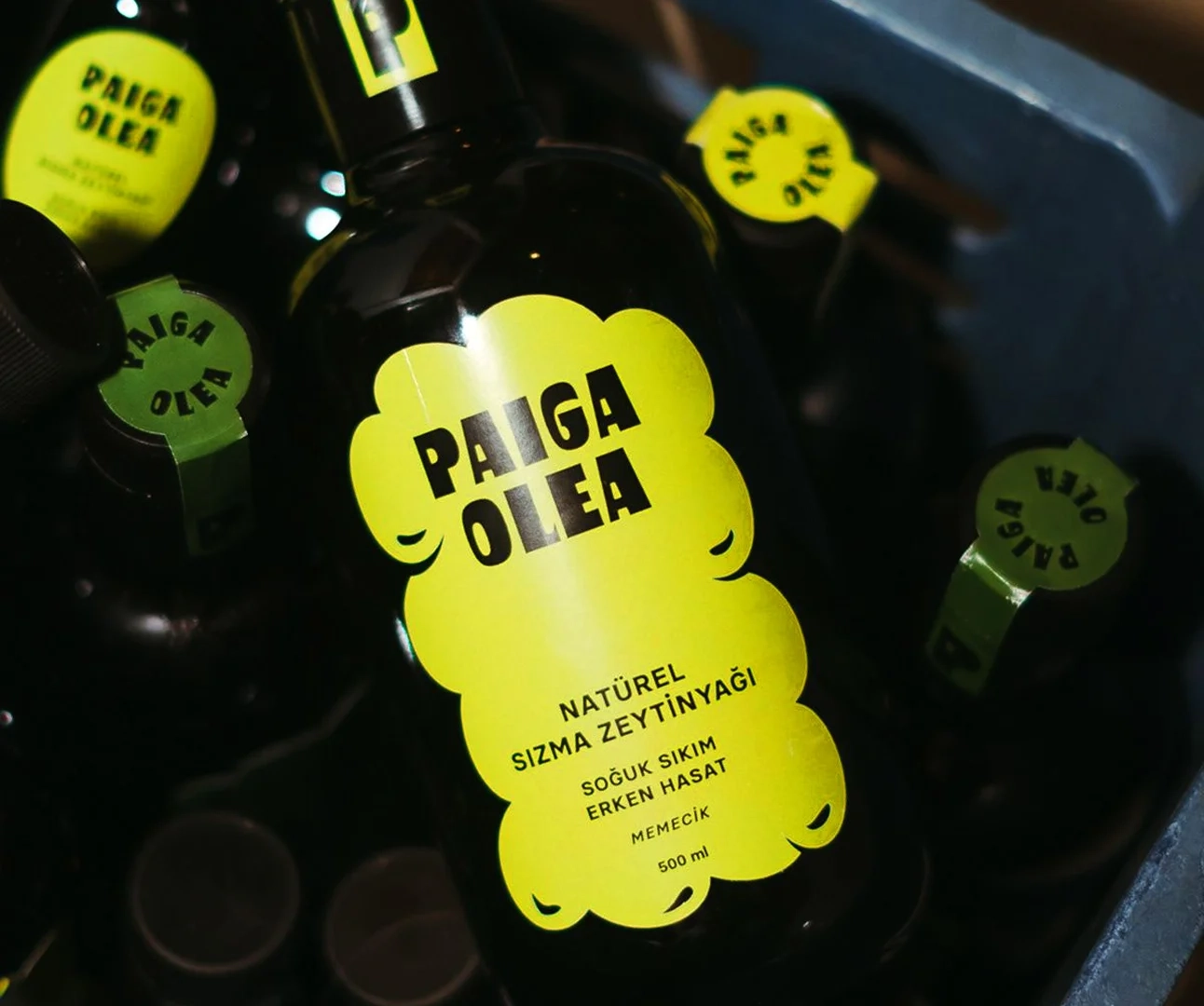 Close-up of a dark glass bottle of Paiga Olea extra virgin olive oil featuring a bright yellow cloud-shaped label with bold black typography and matching green tamper-evident neck seals.
