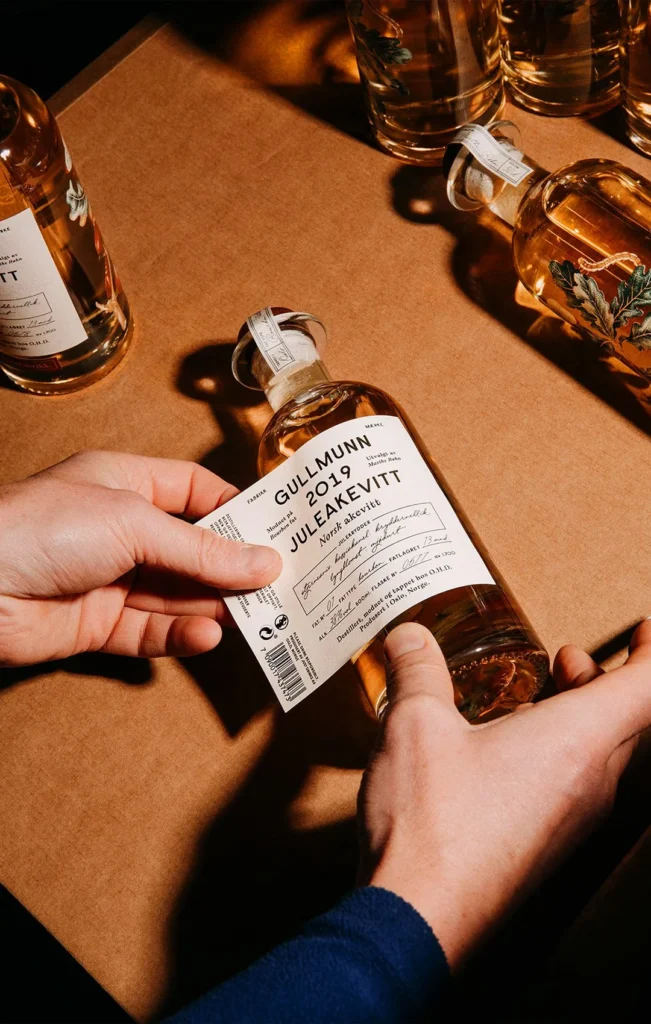Close-up of hands carefully applying a premium white textured paper label to a glass bottle of Gullmunn 2019 Juleakevitt, featuring elegant black typography and a hand-signed batch number.