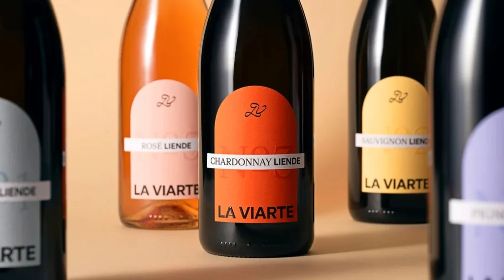 A row of dark glass wine bottles featuring arched minimalist labels in various colors like orange, pink, and yellow, with the brand name "LA VIARTE" and varietals like Chardonnay and Sauvignon printed in clean black typography