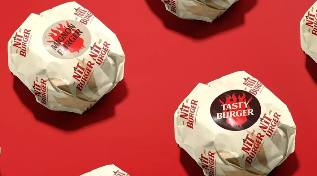 Multiple burgers wrapped in white branded paper, each featuring a large circular sticker with a flame logo and text like "MIGNON BURGER" and "TASTY BURGER" against a bright red background.