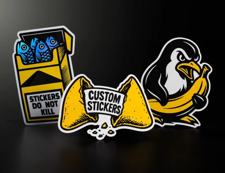 Three custom die-cut vinyl stickers: a yellow cigarette pack parody with blue fish, a broken fortune cookie, and a penguin holding a banana, all with thick white borders
