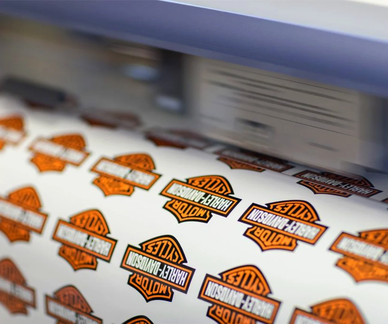 A high-speed digital printer producing a roll of iconic orange, black, and white Harley-Davidson Motor Cycles shield-shaped vinyl stickers