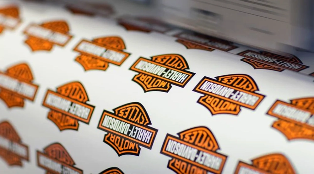 A high-speed digital printer producing a roll of iconic orange, black, and white Harley-Davidson Motor Cycles shield-shaped vinyl stickers.