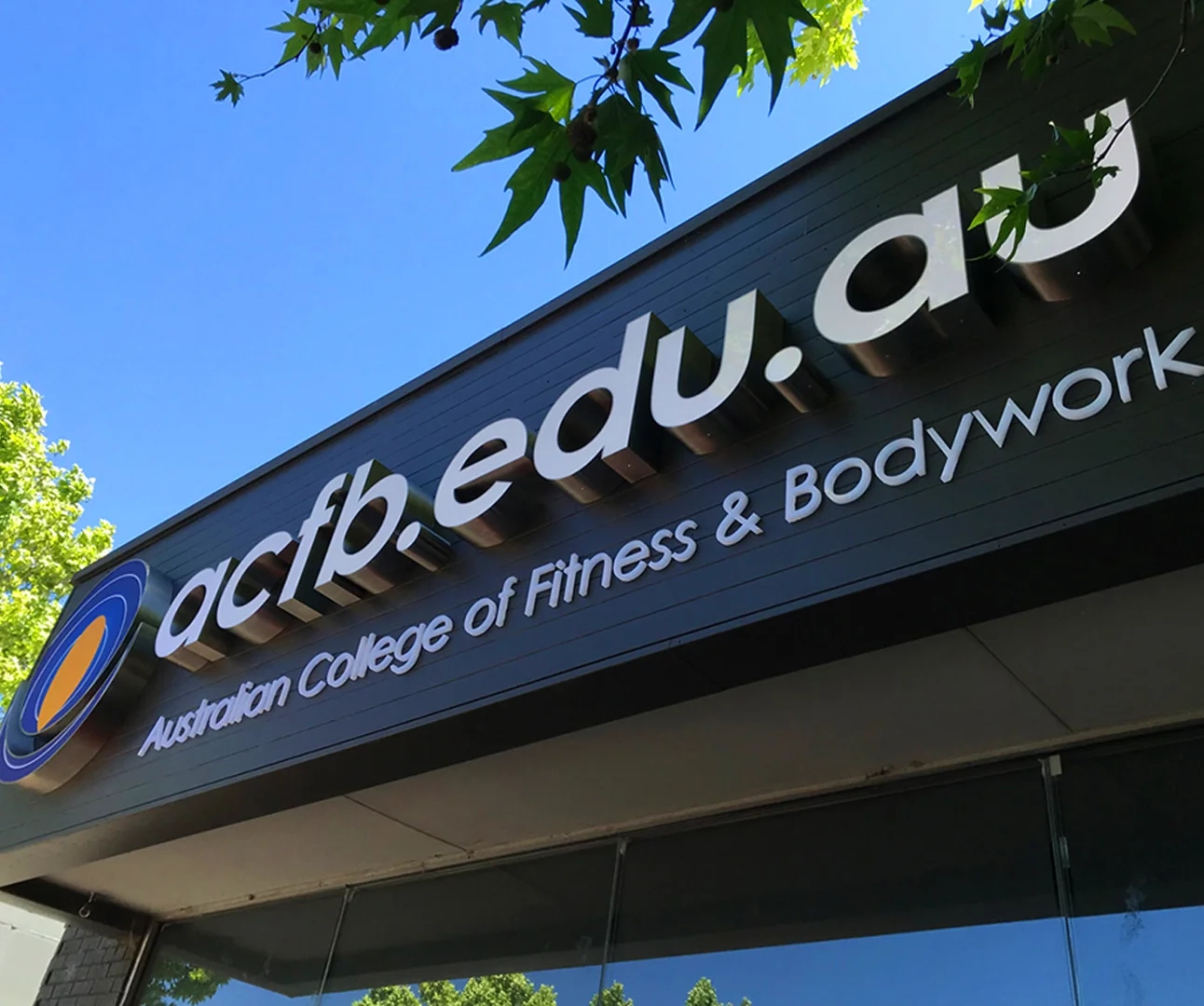 Exterior 3D signage on a black wall for 'acfb.edu.au Australian College of Fitness & Bodywork' under a clear blue sky and green tree branches.