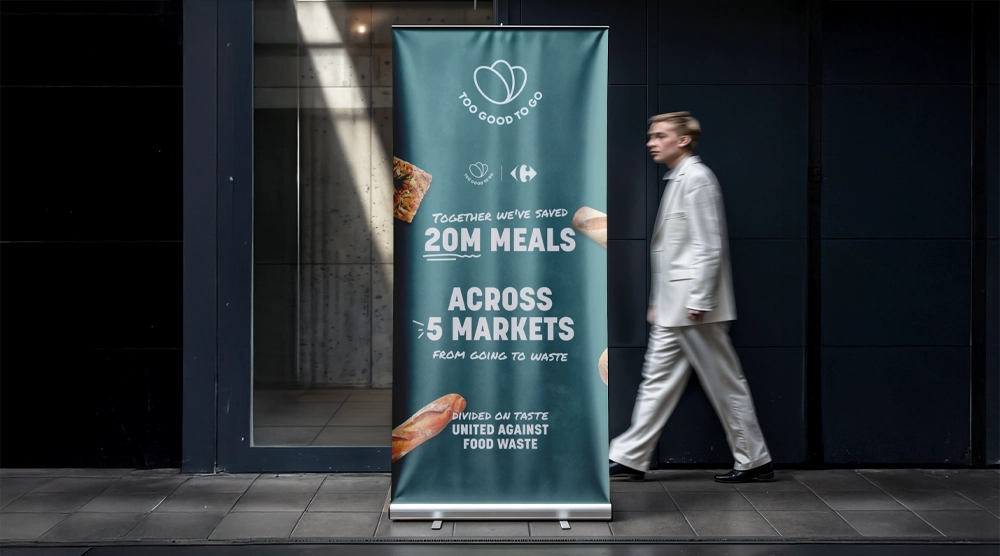A tall teal roller banner for 'Too Good To Go' and 'Carrefour' standing in a modern doorway, featuring bread illustrations and the text 'Together we've saved 20M meals