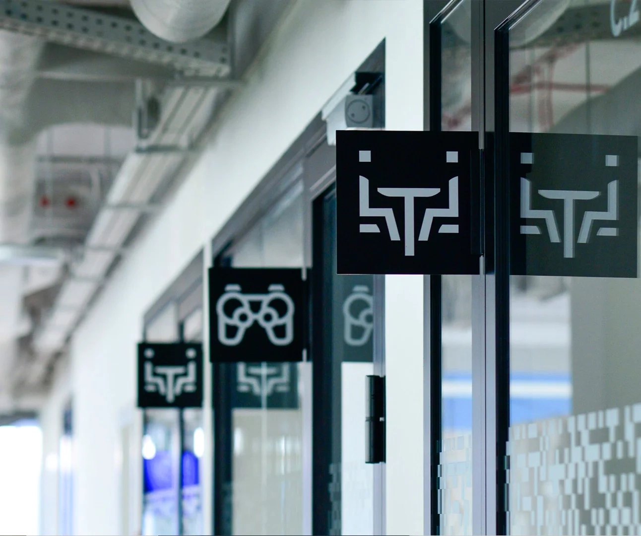 A series of black square signs projecting from glass office doors, featuring white minimalist icons representing a meeting table and chairs, and a game controller