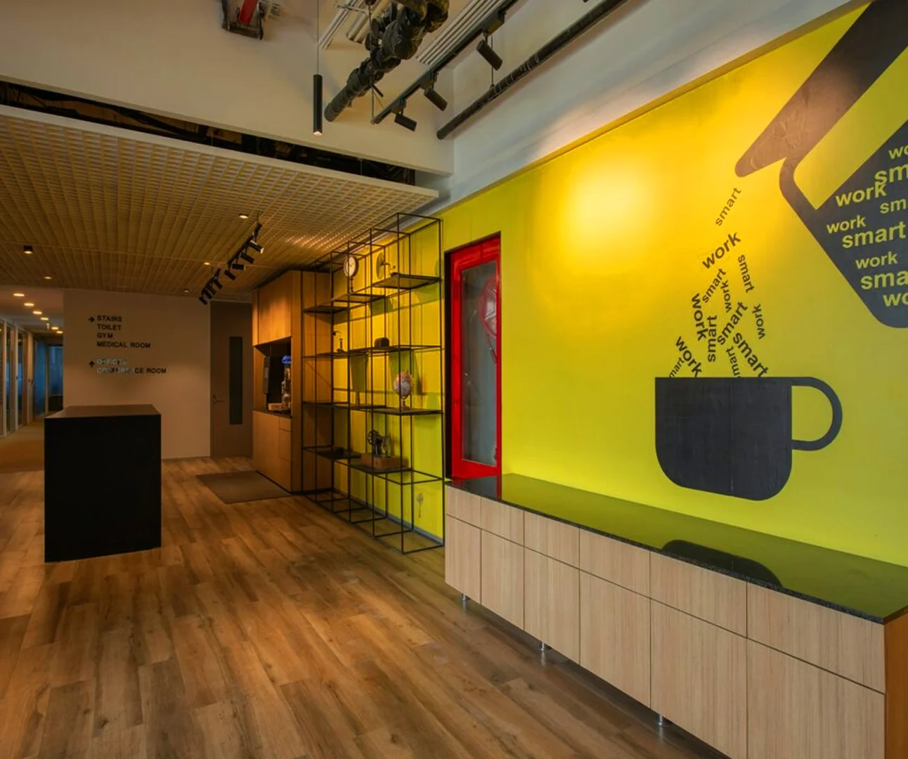 A modern office common area with a vibrant yellow wall featuring a black coffee cup mural with typography, wood flooring, and industrial shelving.