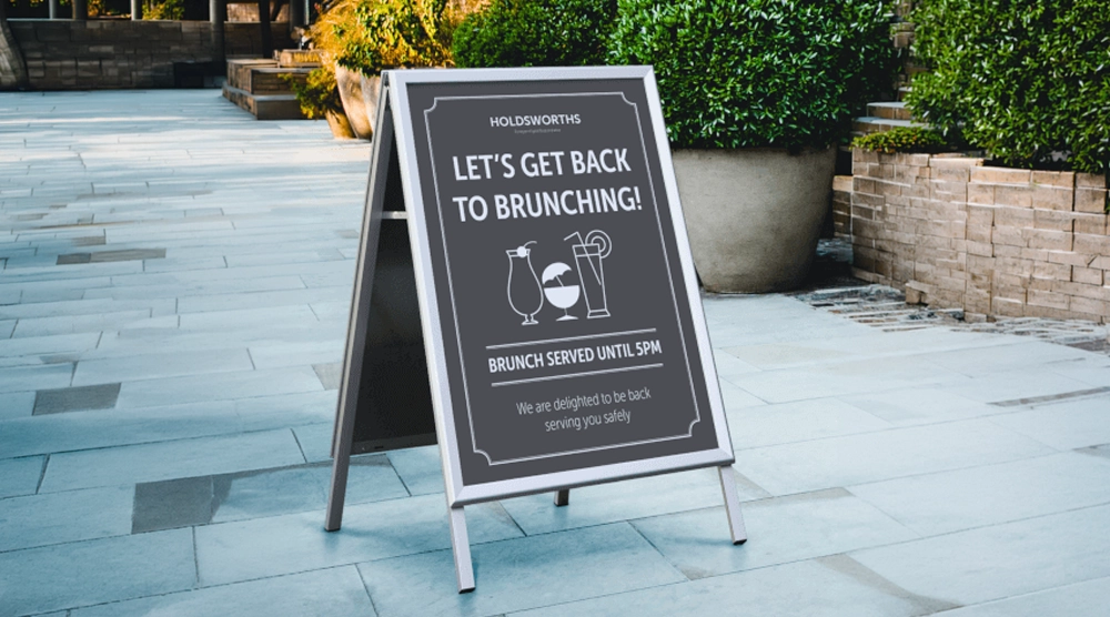 A silver-framed metal A-board pavement sign on a stone walkway featuring a grey poster with cocktail icons and the text "LET'S GET BACK TO BRUNCHING!