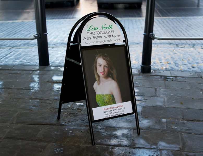A black arched A-board pavement sign on a wet cobblestone street, featuring a professional portrait and a semicircular header with contact information.