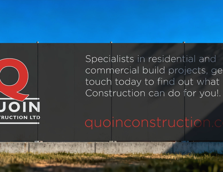 A large black perimeter fence (hoarding) featuring a red and white logo and white promotional text for a construction company against a clear blue sky