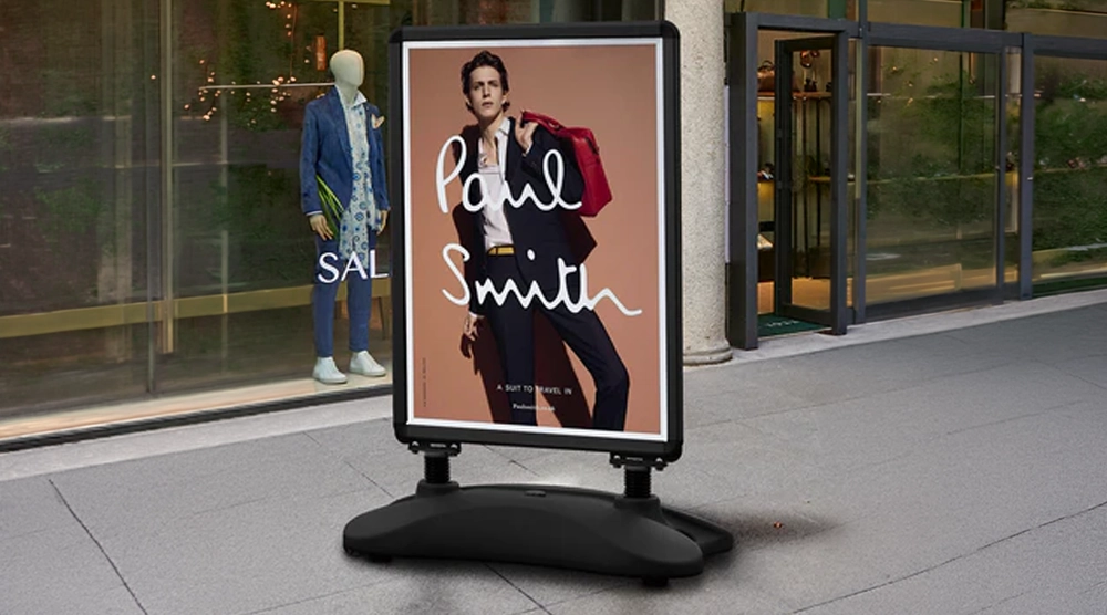 A black-framed pavement swing sign with a heavy-duty base on a sidewalk, featuring a Paul Smith advertisement of a man in a suit carrying a red bag.