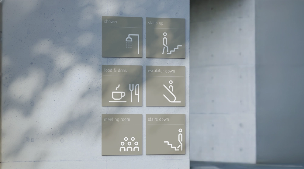 Six square, olive-grey directional signs with white line-art icons and lowercase text mounted on a light grey concrete wall.