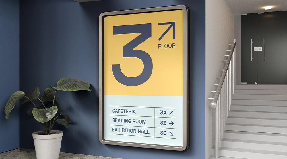 A large, rounded-corner rectangular sign mounted on a blue wall next to a staircase, displaying a large "3" on a yellow background and a directory for a cafeteria, reading room, and exhibition hall on a white background.