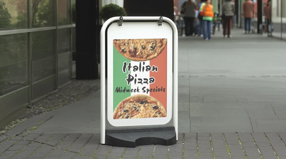 A white-framed swing sign with a black base on a sidewalk, displaying a poster with two pizzas, the Italian flag, and "Italian Pizza Midweek Specials" text.