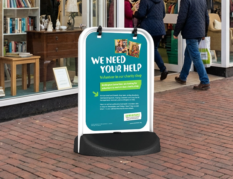 A white-framed outdoor swing sign with a black base on a brick sidewalk, displaying a teal "We Need Your Help" poster for the Samaritans charity shop.