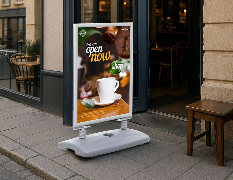A silver-framed pavement swing sign on a sidewalk outside a cafe, featuring a coffee cup graphic and "We are open now" text.