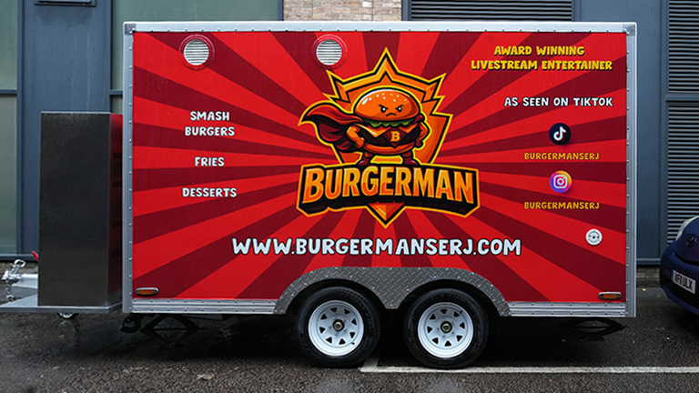 A red and yellow superhero-themed food trailer featuring "Burgerman" branding and social media icons, demonstrating bold trailer wrapping in London.