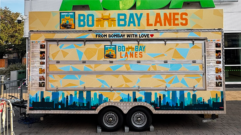 A large dual-axle food trailer wrapped in a yellow geometric pattern with a blue London skyline silhouette, illustrating premium trailer wrapping in London.